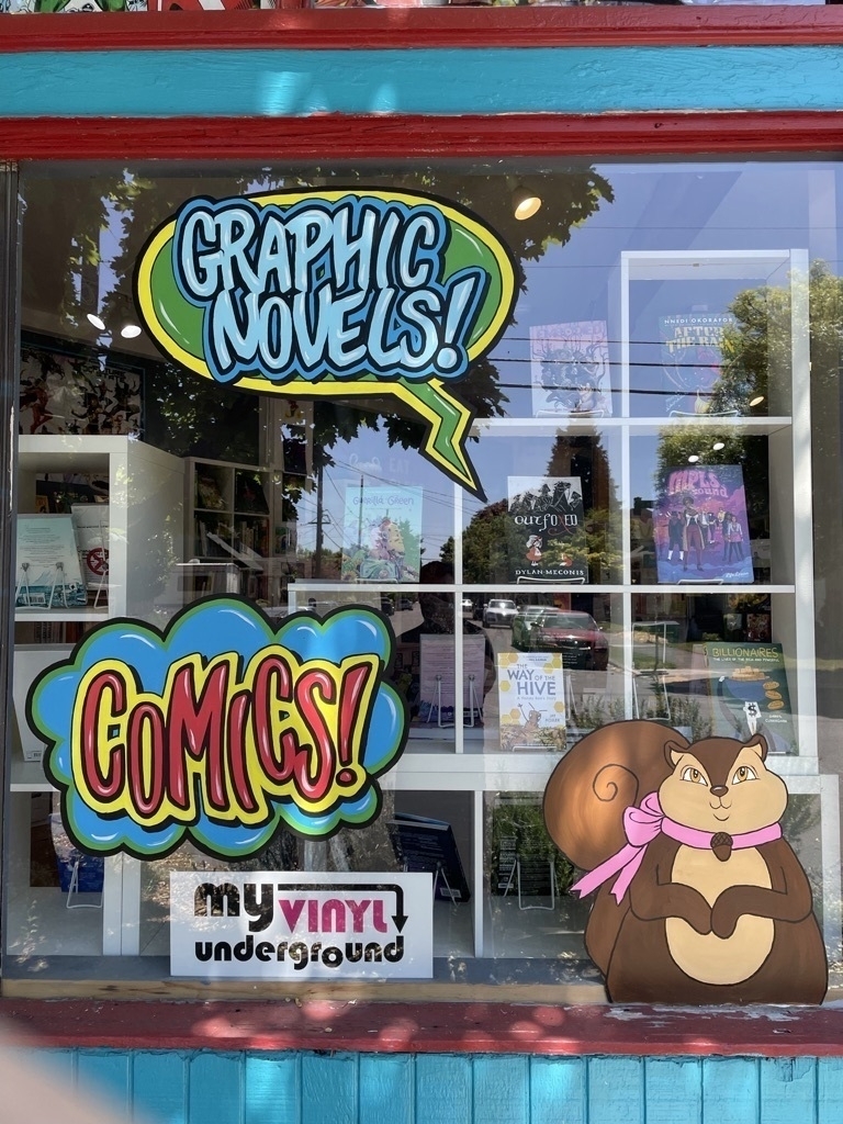 A window display for a comic book store. There are large colorful word baloons that read “Graphic Novels!” and “Comics” painted on the window as well as a large cute cartoon squirrel. 